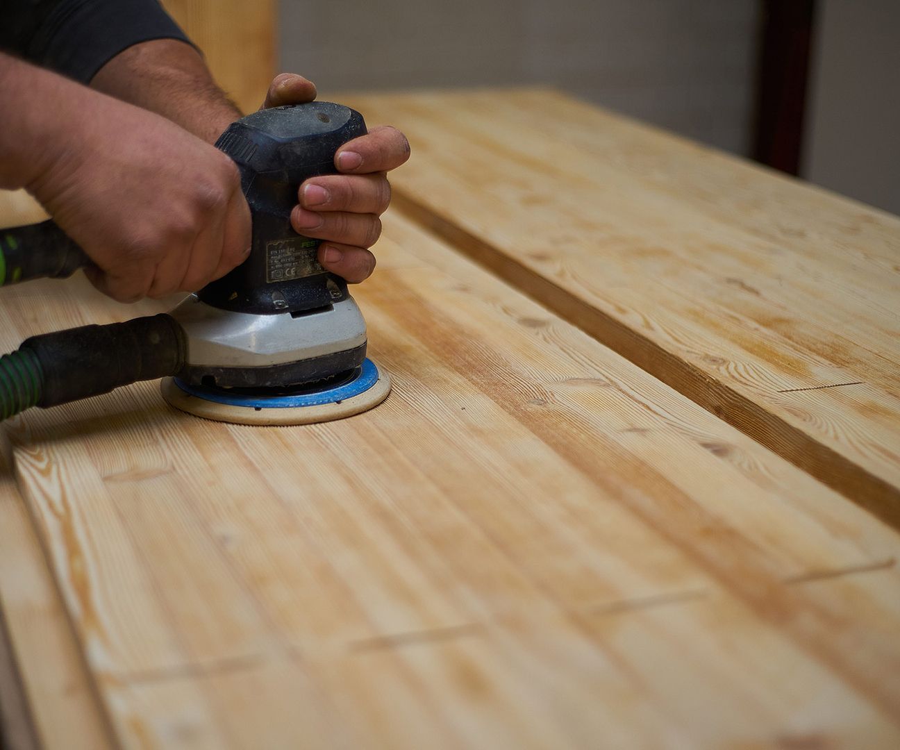 Eine Person schleift einen Holztisch mit einem Schleifgerät, um die Oberfläche zu glätten und vorzubereiten.